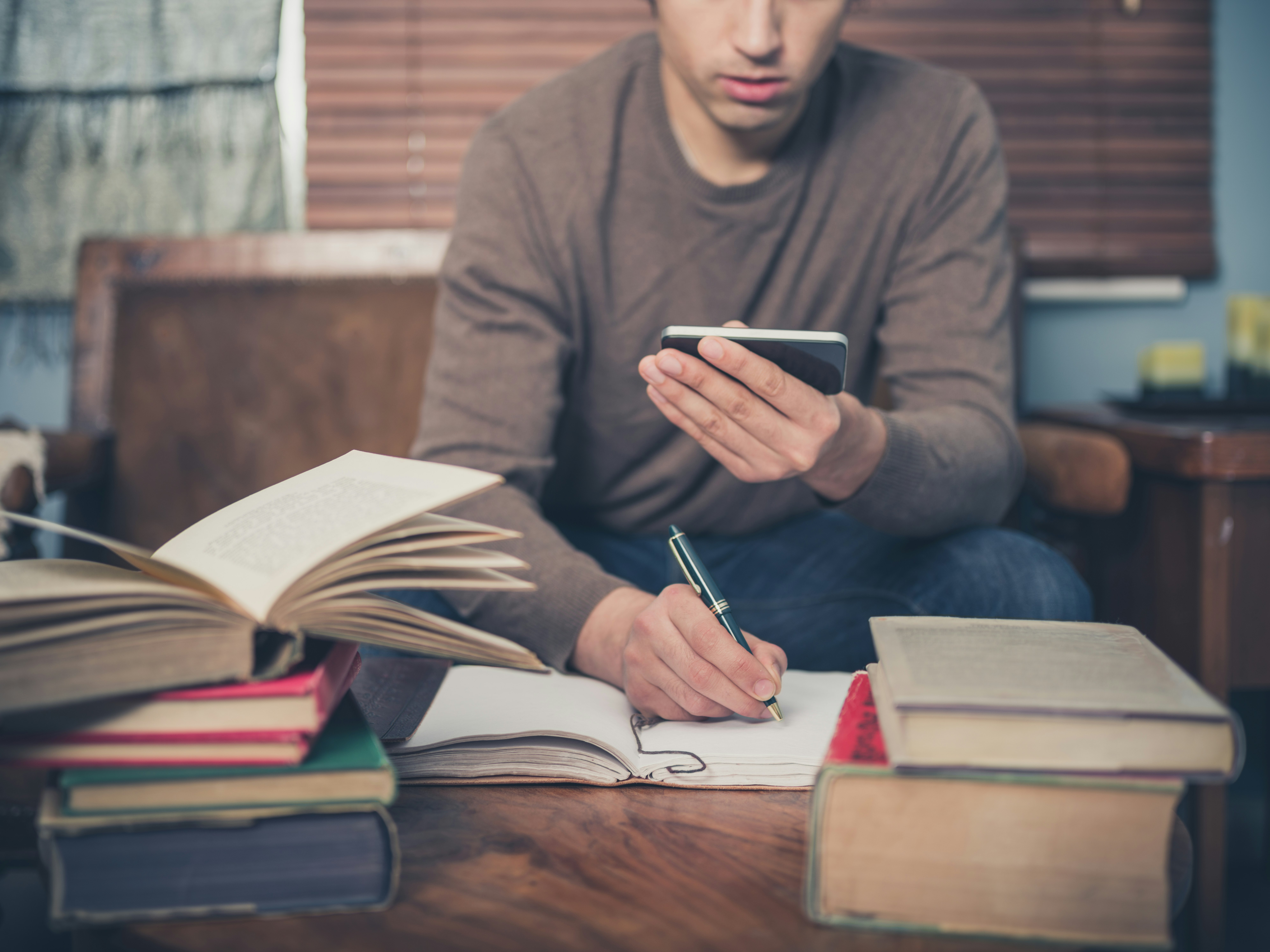 A young man is sitting on a sofa surrounded by books and is using his smartphone